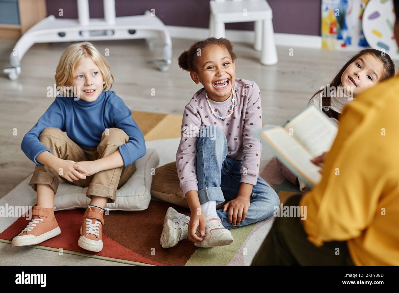 Groupe de petits élèves mignons de l'école maternelle assis sur le plancher de la salle de classe tout en écoutant le professeur les lisant livre Banque D'Images