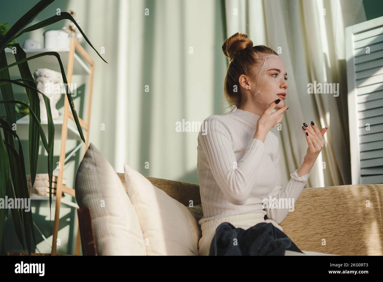 Jeune femme aux cheveux rouges faisant un masque facial, assise sur le canapé à la maison. Soins médicaux. Femme visage soins de la peau cosmétique. Traitement thérapeutique Banque D'Images