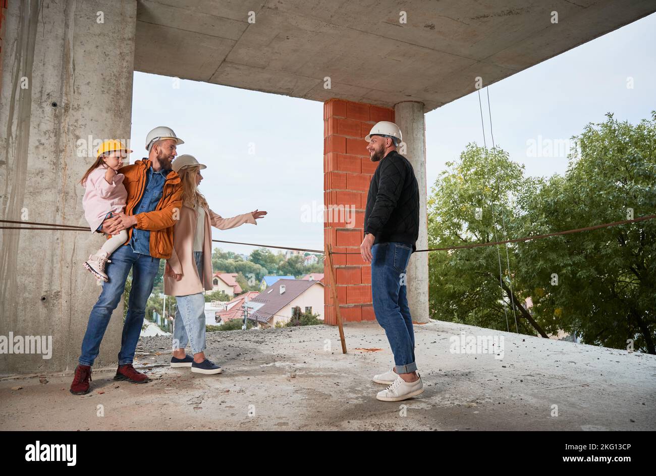 Homme et femme avec enfant debout à l'intérieur de l'immeuble d'appartements en construction et parlant avec le constructeur. Famille futurs propriétaires discutant du processus de construction avec le travailleur de la construction. Banque D'Images