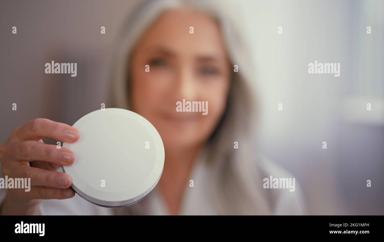 Vieille caucasienne 50s femme mature senior grand-mère dame avec de longs cheveux gris dans peignoir montrant un pot de crème minérale naturelle de crème solaire biologique avec Banque D'Images