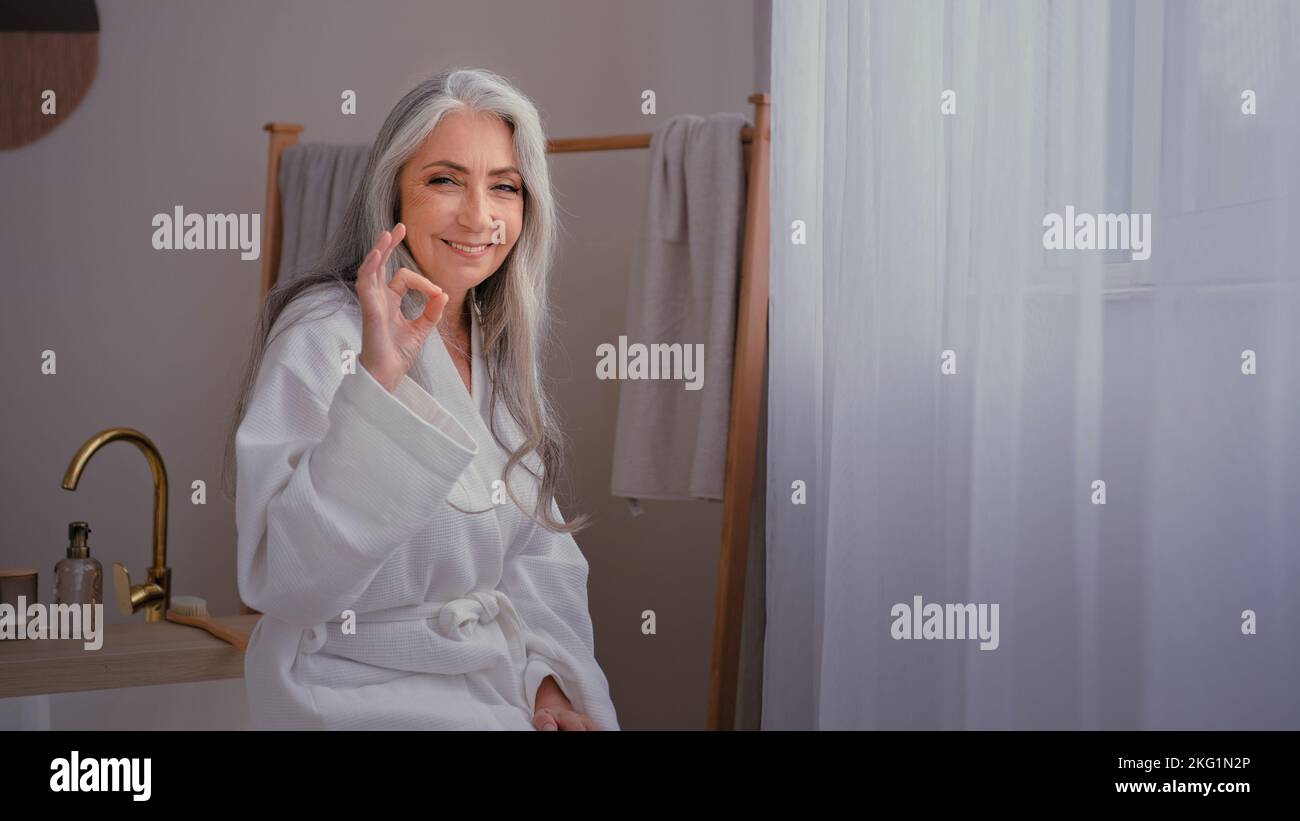 Femme âgée mature femme caucasienne ancienne femme à cheveux gris femme dans peignoir dans la salle de bains après avoir lavé la procédure de beauté montrant ok recommander geste Banque D'Images