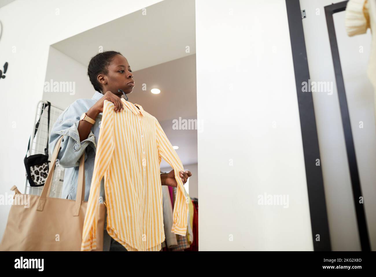 Portrait à faible angle d'une jeune femme noire essayant de porter des vêtements par miroir tout en faisant des achats durables dans un magasin d'épargne Banque D'Images