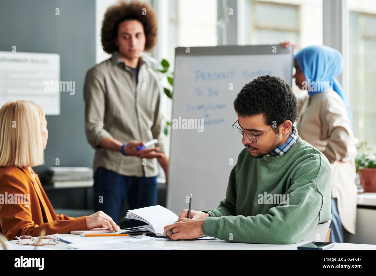 Jeune homme faisant des notes en copybook tout en étant assis par bureau à la leçon de grammaire anglaise contre un gars expliquant le nouveau sujet aux femmes Banque D'Images