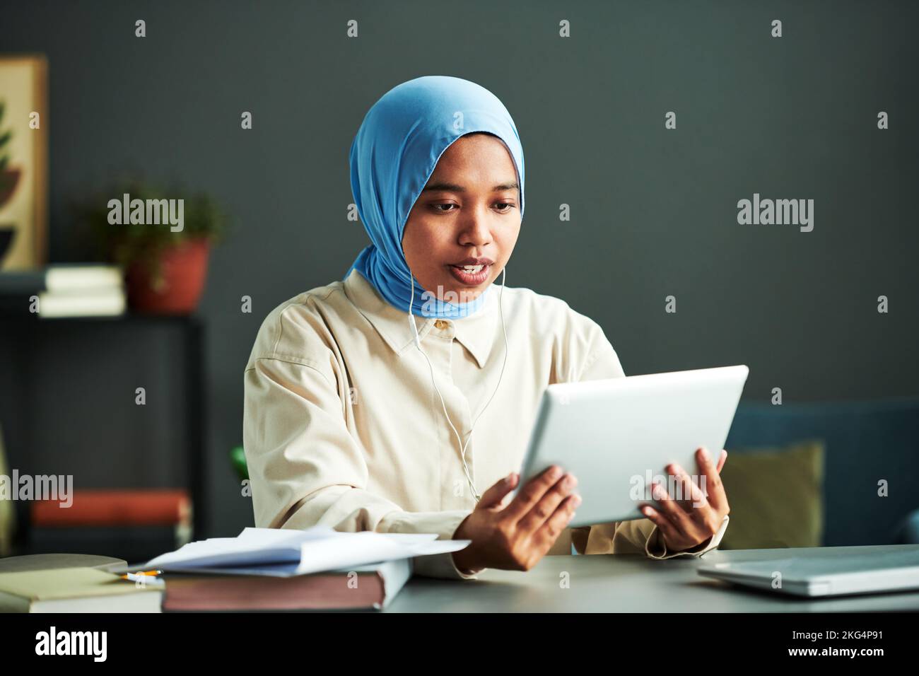 Jeune femme musulmane confiante dans les écouteurs communiquant avec le public en ligne tout en regardant l'écran de la tablette pendant la leçon Banque D'Images