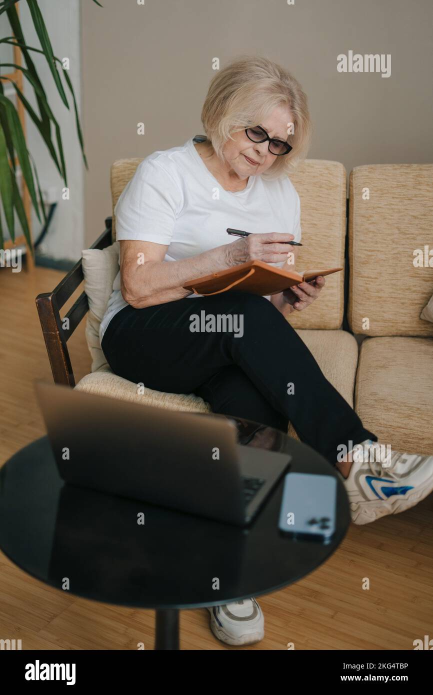 Portrait d'une femme âgée aux cheveux gris, assise sur un canapé, prenant des notes dans un bloc-notes à la maison avec un ordinateur portable ouvert. Communication Internet. Éducation Banque D'Images