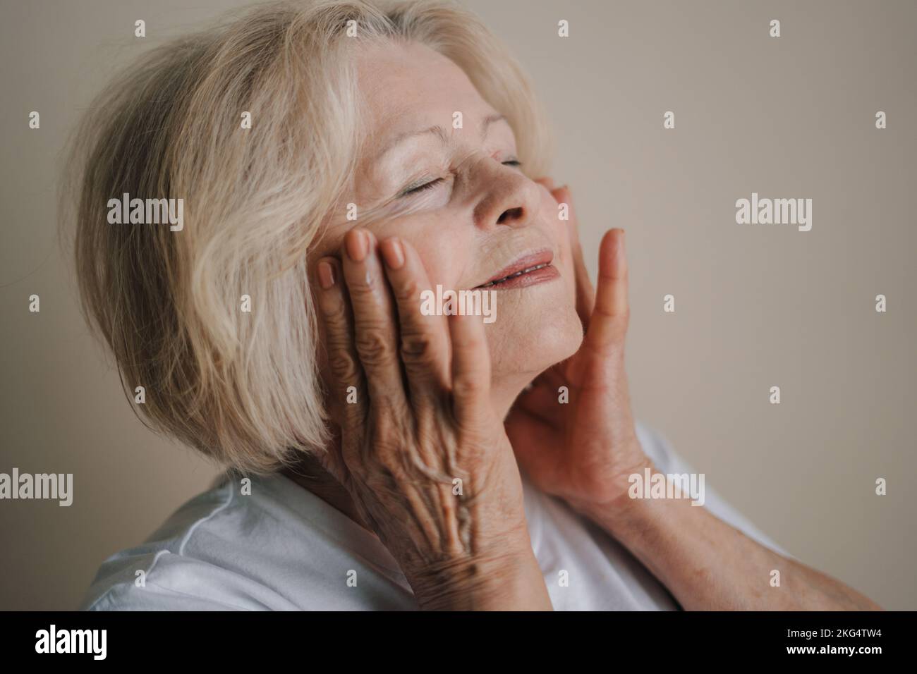 Portrait d'une belle femme âgée souriante touchant sa joue, après avoir utilisé des cosmétiques anti-âge le matin isolés sur fond gris. Personnes Banque D'Images