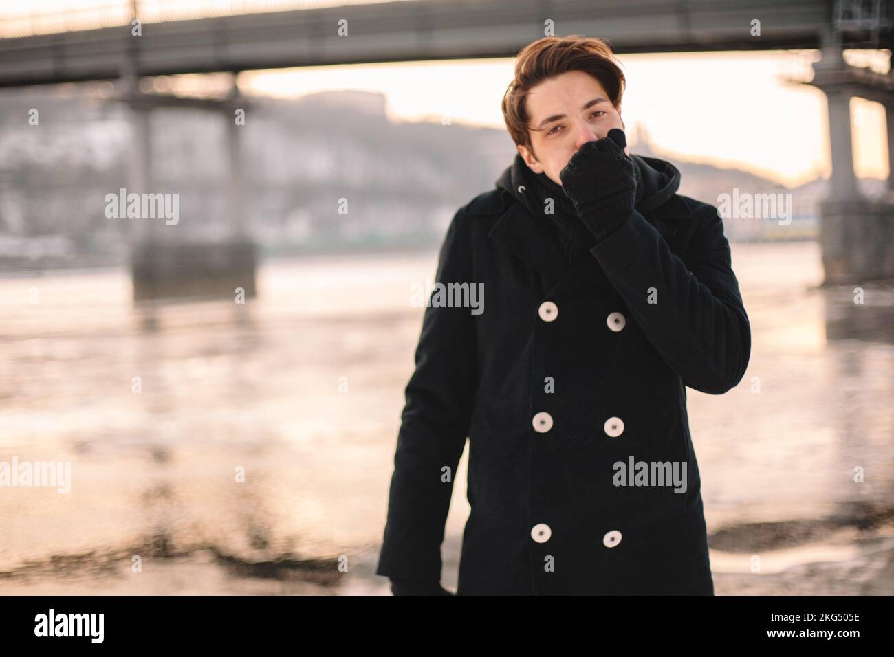 Jeune homme se réchauffant les mains tout en se sentant debout au bord de la rivière par temps gelé en hiver Banque D'Images