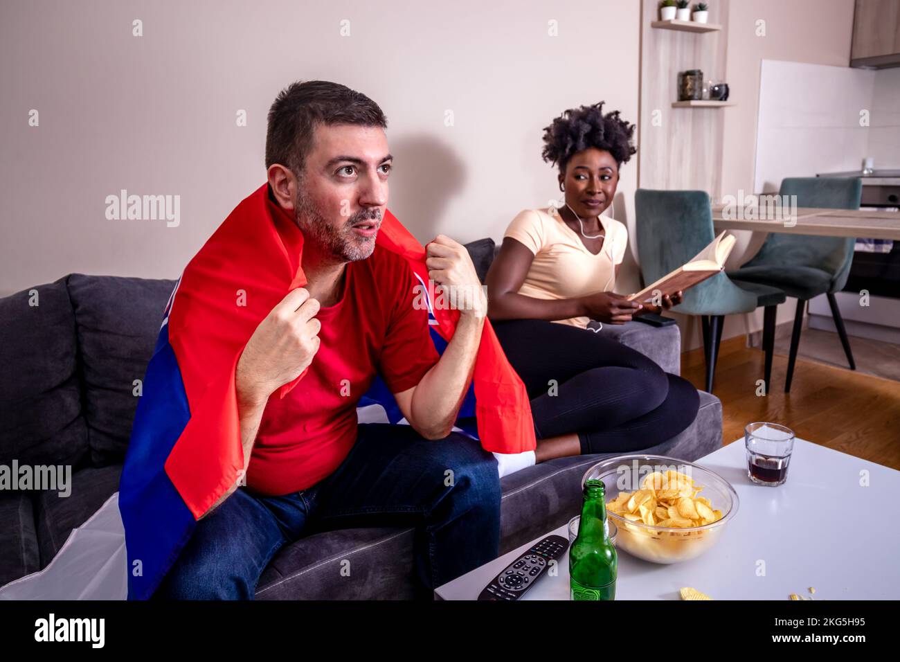 Le mari regarde un match de football à la télévision, tandis que sa femme s'ennuie. La femme essaie de lire un livre avec des écouteurs dans ses oreilles. Coupe du monde. Banque D'Images