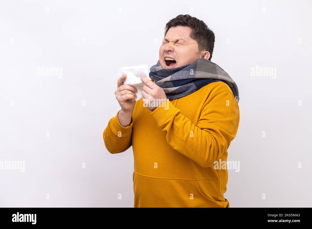 Homme enveloppé d'un foulard éternuant dans une serviette, nettoyant le nez et la toux, souffrant de symptômes de la grippe, fièvre, portant un sweat à capuche de style urbain. Studio d'intérieur isolé sur fond blanc. Banque D'Images