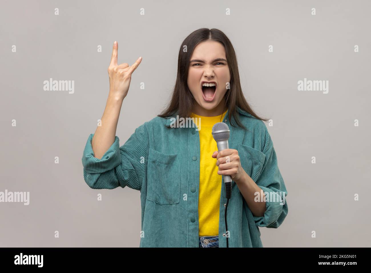 Portrait d'une femme attrayante avec des cheveux sombres chantant des chansons préférées avec un microphone et montrant le rock and roll geste, en portant une veste de style décontracté. Prise de vue en studio isolée sur fond gris. Banque D'Images