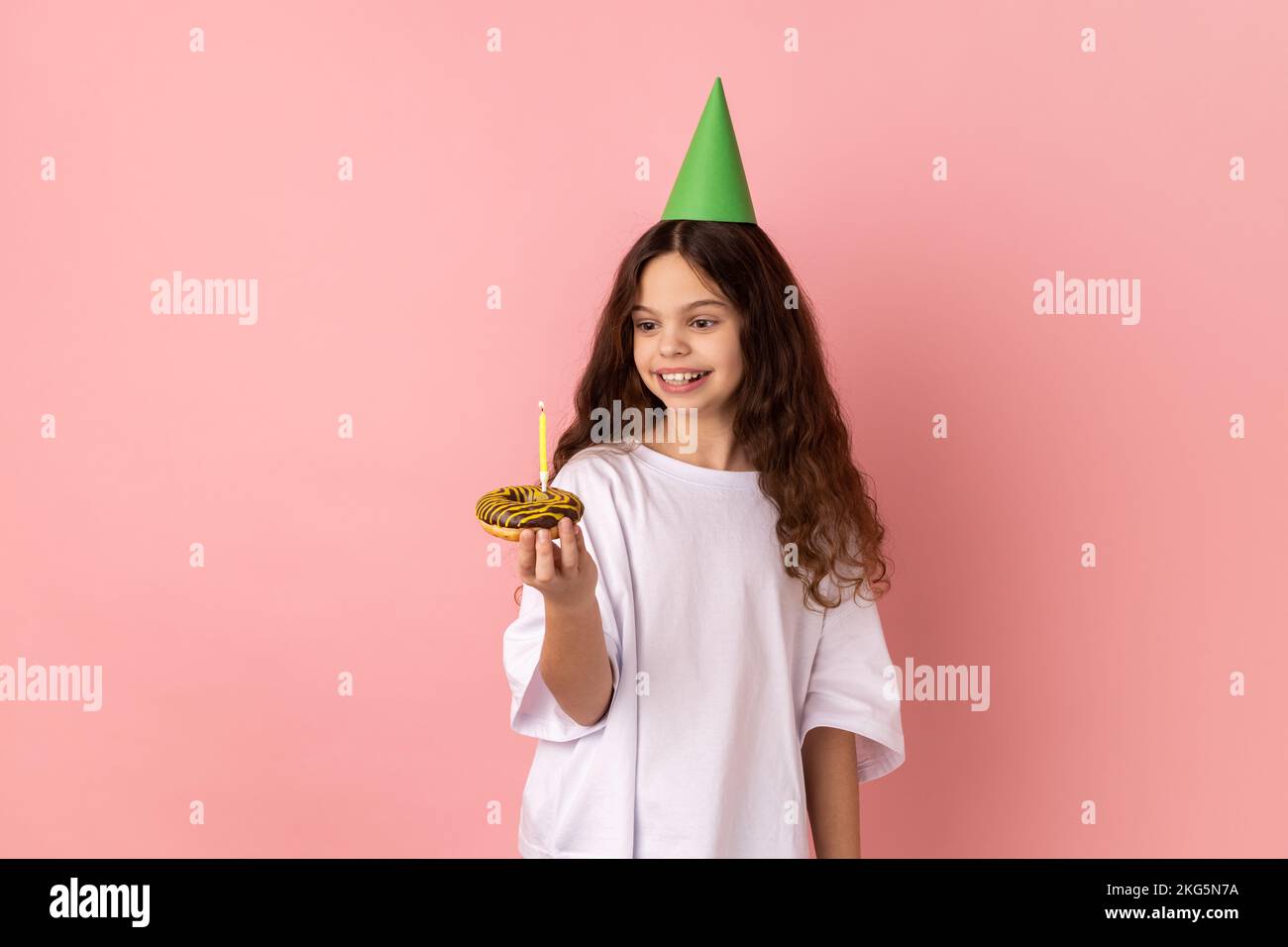 Une petite fille satisfaite en forme de cône vert sur la tête tenant un gâteau avec une bougie et regardant le dessert avec un sourire savoureux, exprimant le bonheur pur de l'enfant. Studio d'intérieur isolé sur fond rose. Banque D'Images