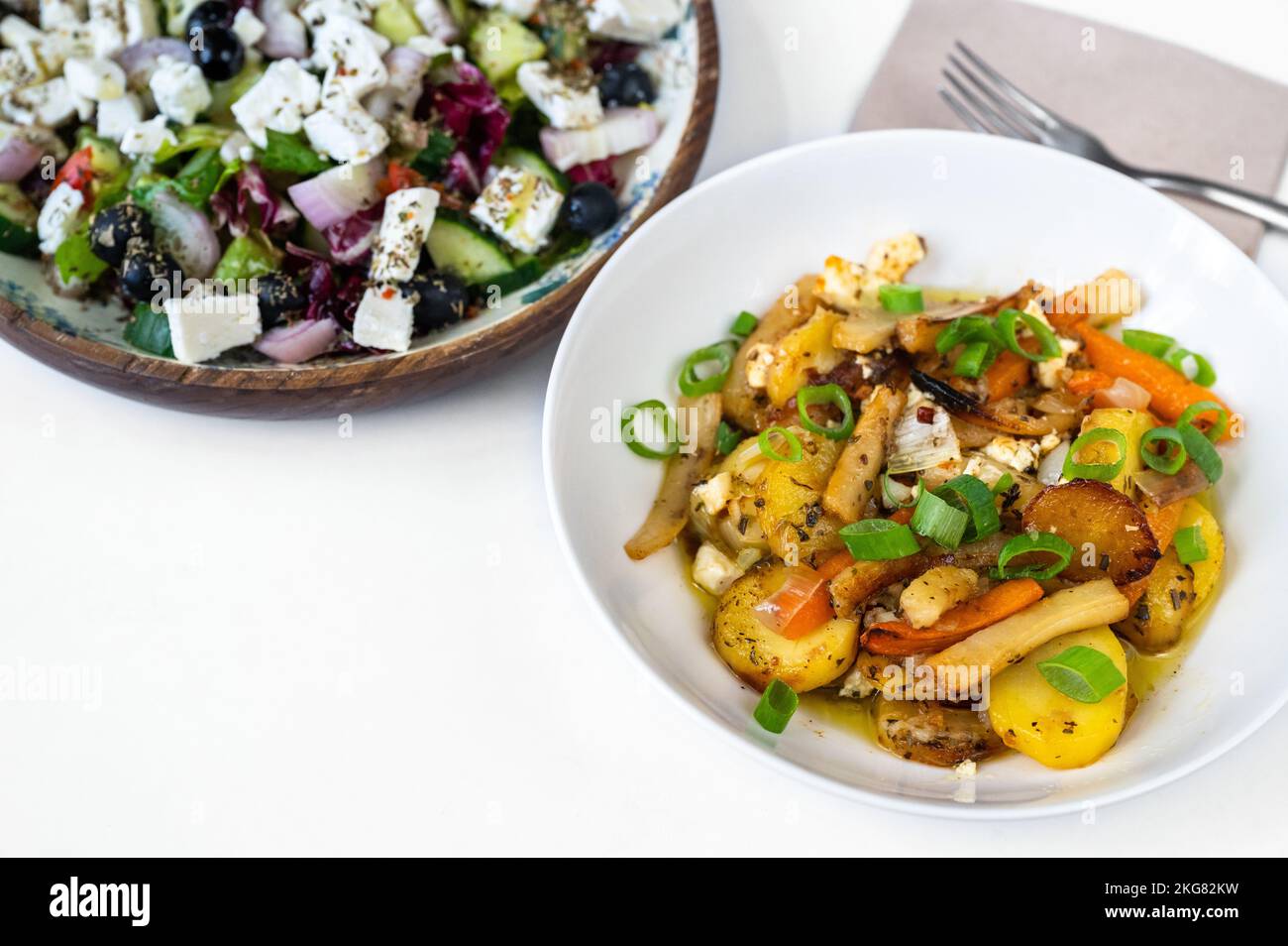 Salade grecque dans une partie de la première assiette, pommes de terre frites, carottes, oignons, ail et persil dans la deuxième assiette sur fond blanc. Déjeuner végétarien sain ou din Banque D'Images