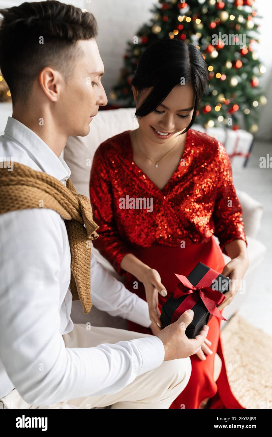 Un jeune mari offre un cadeau de Noël à une femme asiatique enceinte sur un canapé à la maison Banque D'Images