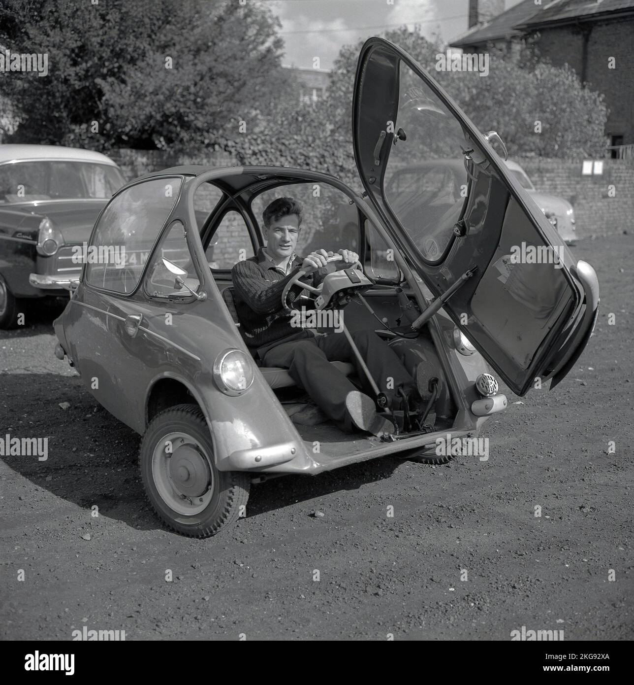 1960s, historique, à l'extérieur dans un parking en gravier, un homme assis à l'intérieur d'une petite voiture de l'époque, une voiture à bulles, une BMW Isetta, qui avait une porte d'ouverture avant, Wycombe, Angleterre, Royaume-Uni. Construite pour la première fois par ISO spa en Italie en 1953, cette petite voiture a été construite sous licence dans différents pays, y compris par BMW en Allemagne jusqu'en 1962 et connue sous le nom de « Bubble car ». Banque D'Images