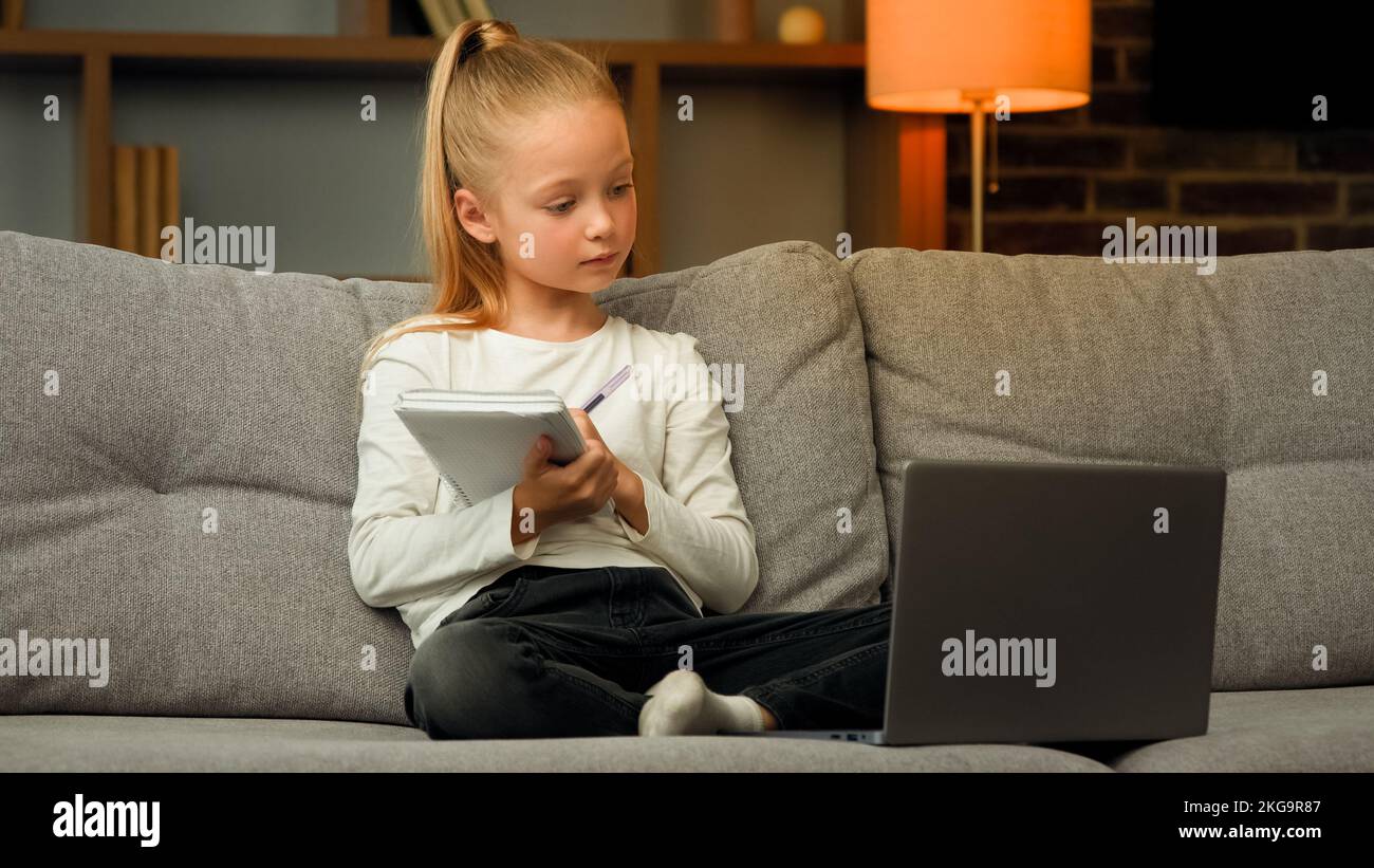 Blonde petite fille intelligente élève d'école écouter la leçon éducative sur ordinateur portable à la maison en ligne réunion de classe dans le chat vidéo pendant la pandémie écrire des devoirs Banque D'Images