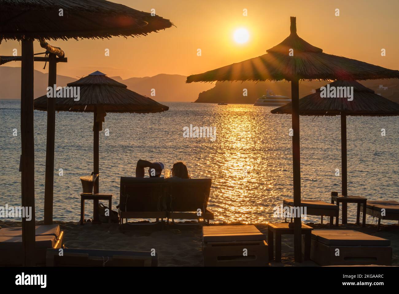 IOS, Grèce - 13 septembre 2022 : vue d'un couple allongé sur des chaises longues, boire du vin et profiter de l'incroyable coucher de soleil orange dans iOS Grèce Banque D'Images