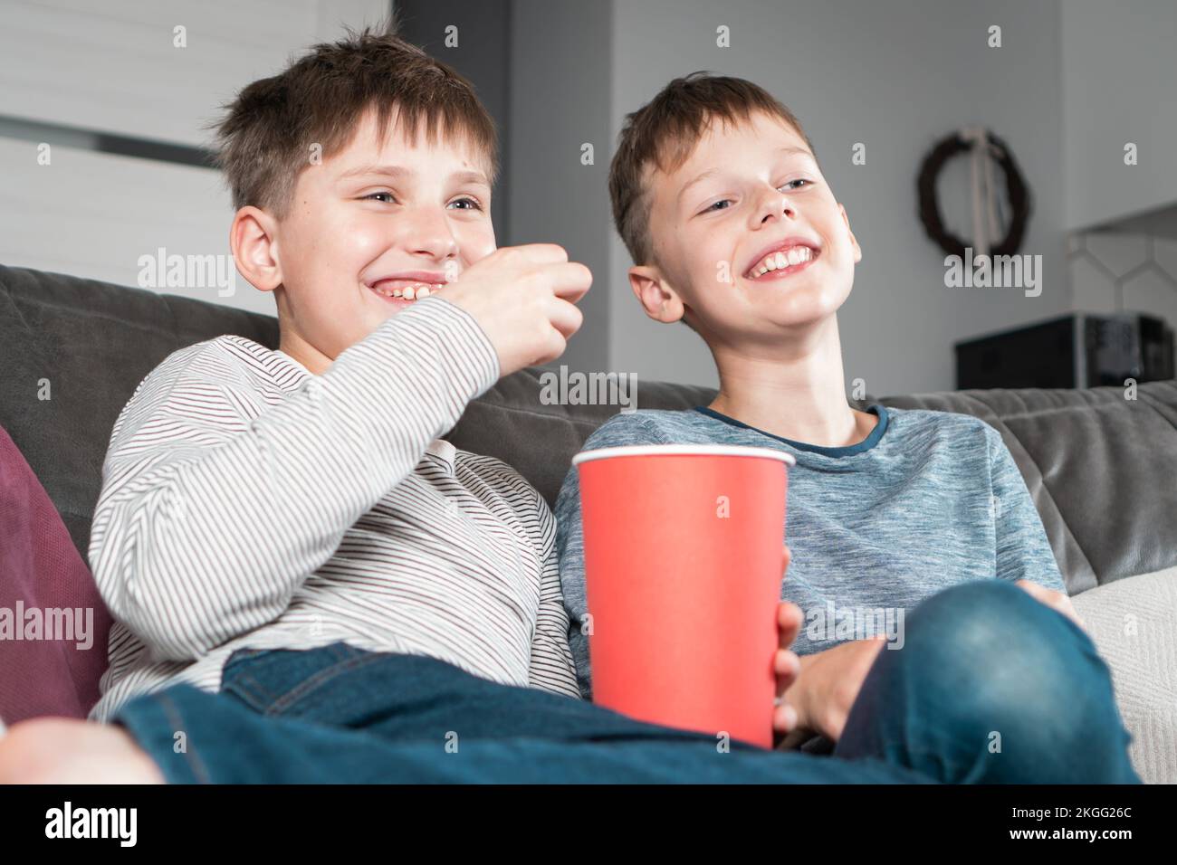 Portrait de deux adolescents qui rient positivement assis sur un canapé à la maison, regardant un film humoristique vidéo à la télévision, en mangeant du pop-corn rouge Banque D'Images