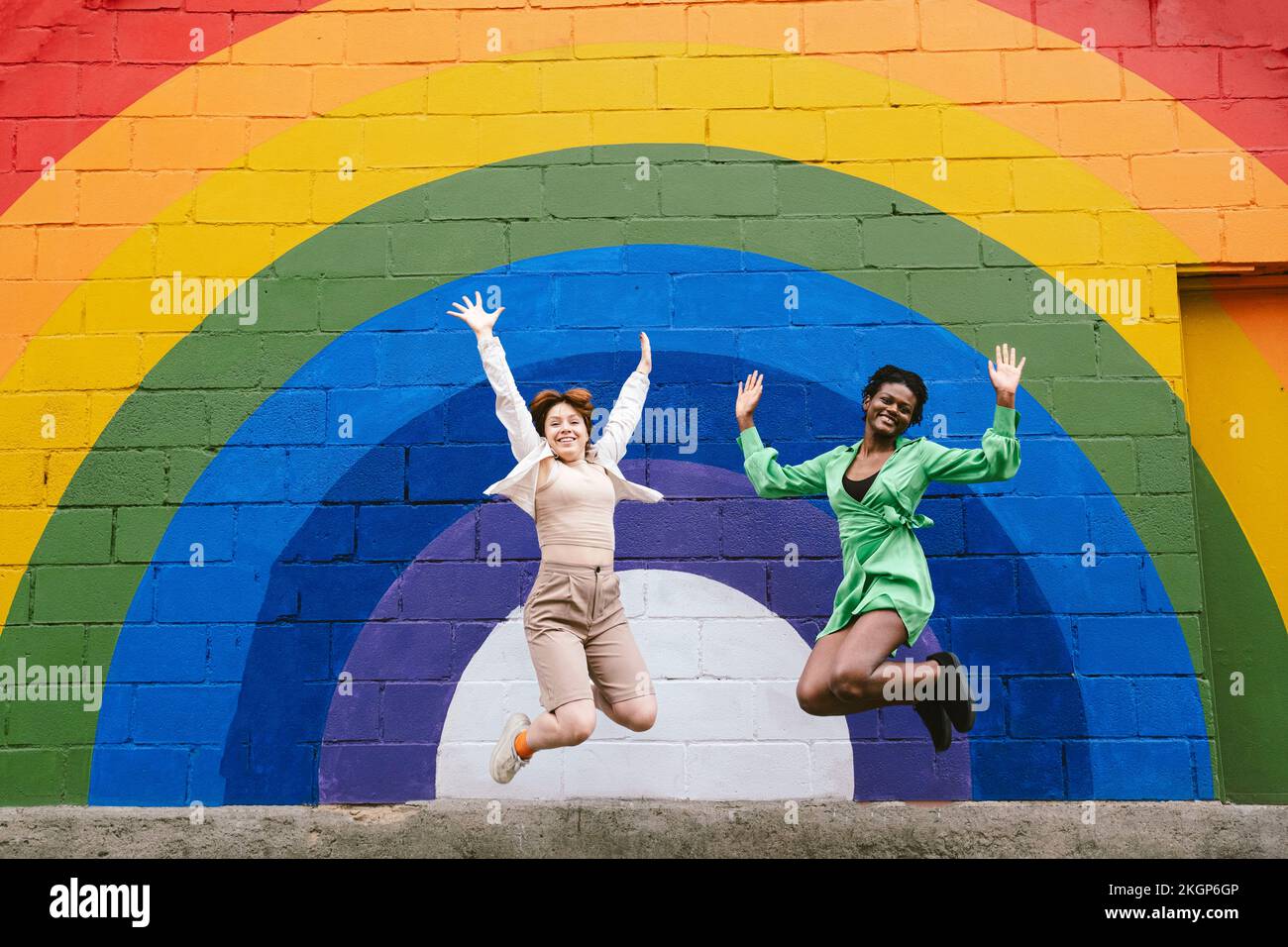 Jeune femme souriante avec un ami qui saute devant le drapeau arc-en-ciel peint sur le mur Banque D'Images