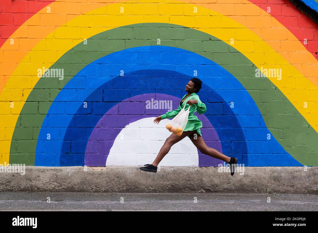 Jeune femme souriante tenant un sac en maille sautant devant le drapeau arc-en-ciel peint sur le mur Banque D'Images