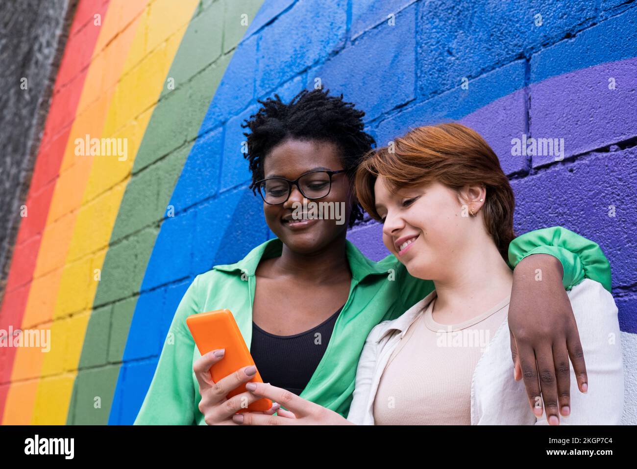 Femme souriante partageant un smartphone avec un ami devant un mur de couleur arc-en-ciel Banque D'Images