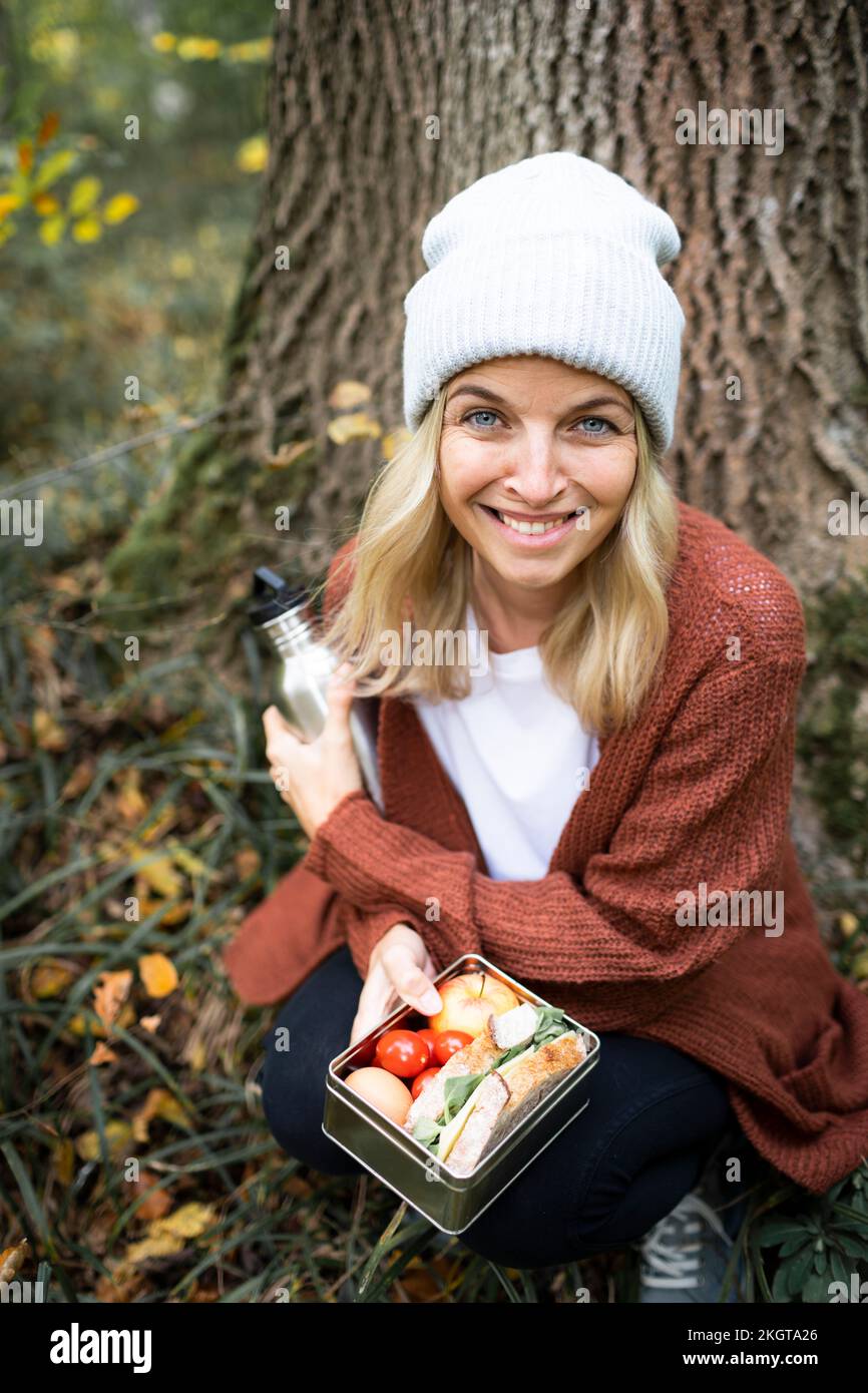 Femme souriante avec boîte à lunch qui s'accroupite dans la forêt le week-end Banque D'Images