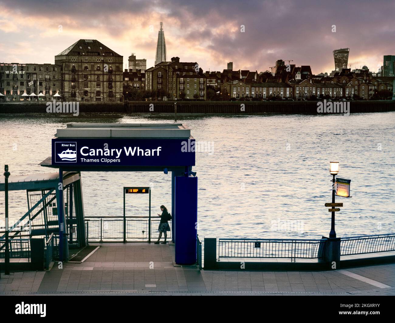 Femme seule utilisant un smartphone au coucher du soleil sur la jetée de Canary Wharf River Boat avec London Shard et le bâtiment Walkie Talkie en arrière-plan Londres E14 Banque D'Images