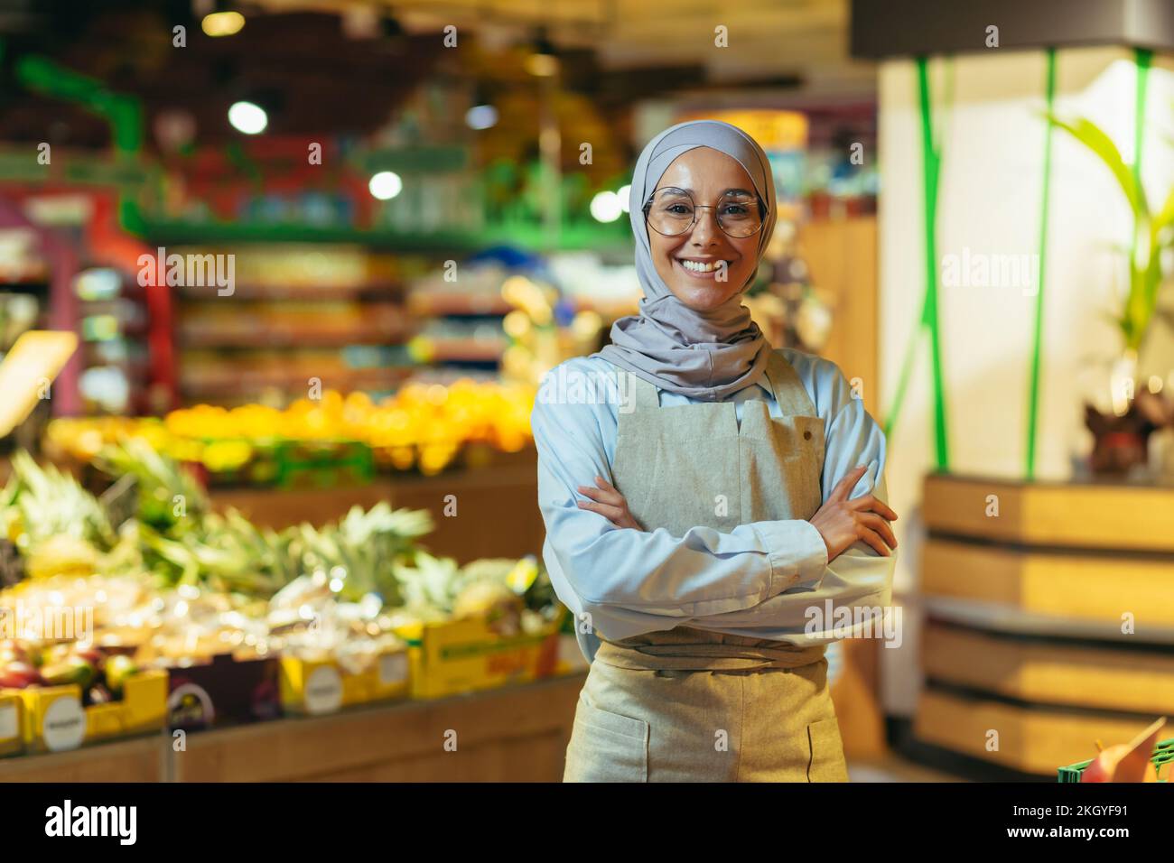 Portrait d'une femme dans un hijab en tant que vendeur dans un supermarché, une vendeuse aux bras croisés sourires et regarde la caméra, un tablier vend des pommes et des fruits, une femme musulmane est satisfaite dans les verres. Banque D'Images