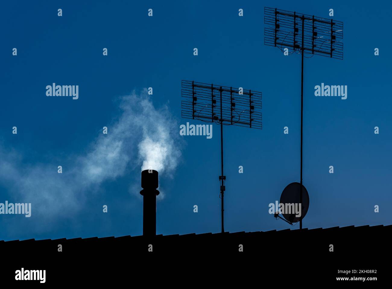 Cheminée avec fumée blanche, antennes tv et parabole satellite vu en silhouette sur le toit, espace de copie Banque D'Images