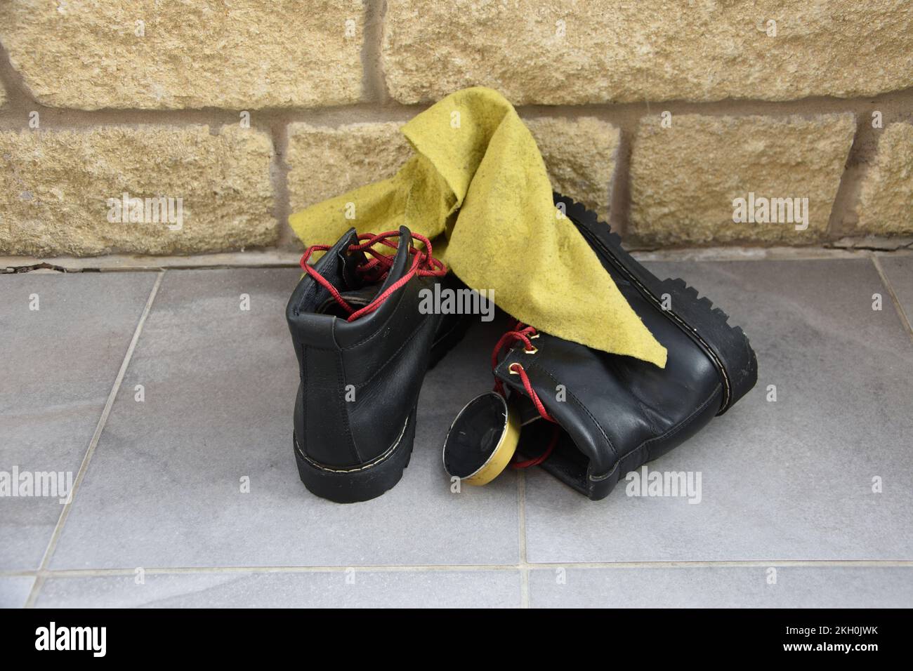 Bottes de marche noires avec lacets rouges attendant le nettoyage sur sol carrelé avec briques de pierre à l'arrière et un plumeau jaune avec une boîte de cirage de chaussures. Après Banque D'Images