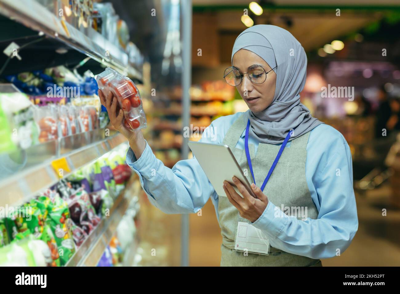 Une femme musulmane dans un hijab, un ouvrier de supermarché vérifie les produits sur les étagères et dans les réfrigérateurs, une femme avec une tablette et un tablier utilise une tablette pour examiner les produits. Banque D'Images