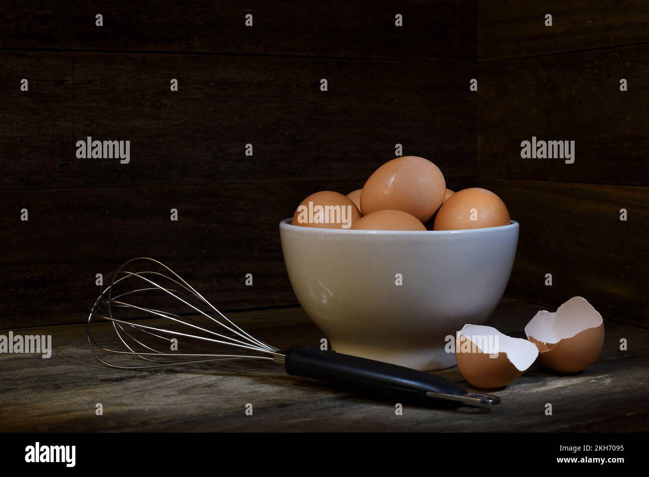 Un bol blanc d'œufs de poulet à portée libre à côté d'un fouet en ballon et de deux coquilles d'œufs dans un cadre doux, sombre, moody et rustique; capturé dans un studio Banque D'Images