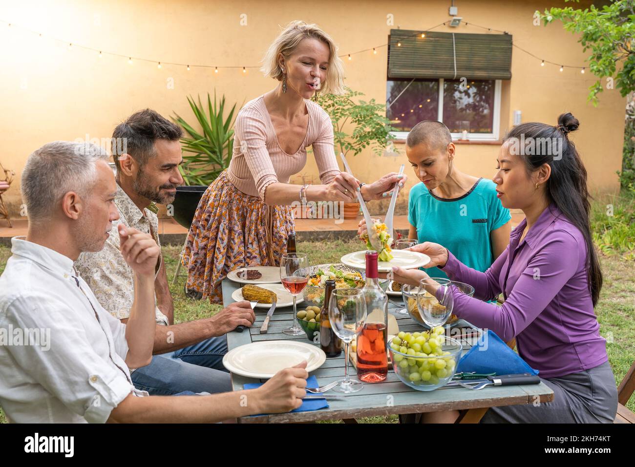 Des amis heureux rient avec un grand sourire autour de la table. La femme sert la nourriture dans le patio de la maison. Sourires, gaieté concept. Banque D'Images