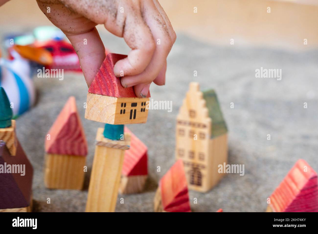 Main de bébé avec de petites maisons de jouets en bois sur le sable. Jeu pour enfants. Cinq cottages avec toit rouge sur sol gris. Village de modélisation. Construction et Banque D'Images