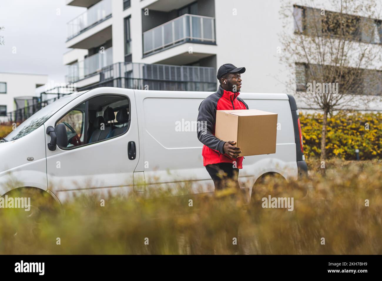 L'utilisation des véhicules dans la livraison des colis. Photo extérieure de beau livreur noir devant son camion blanc qui marche vers la maison du client. Premier plan flou. Photo de haute qualité Banque D'Images