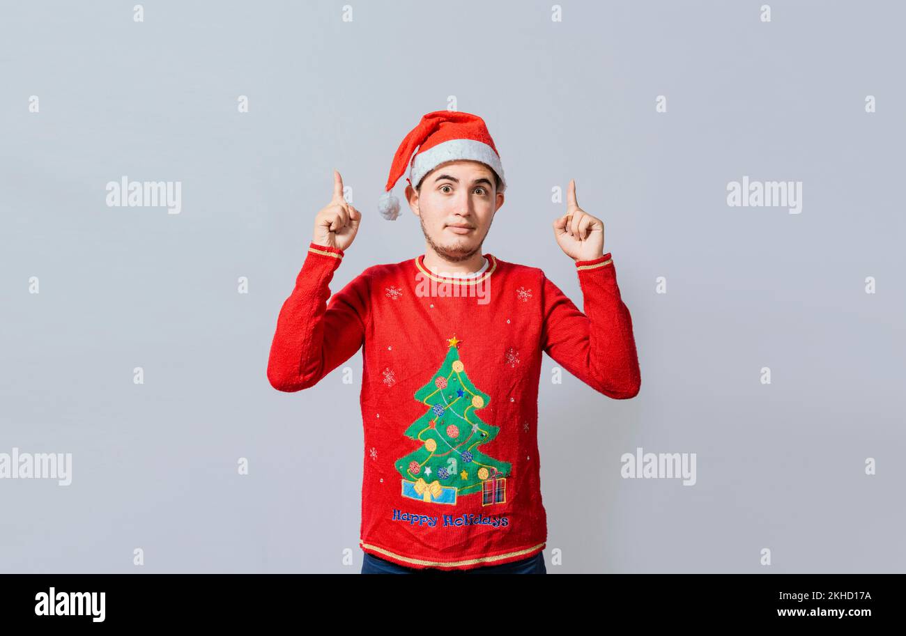Guy dans les vêtements de Noël pointant et recommandant. Portrait d'un gars en chandail et chapeau de noël pointant vers le haut isolé. Jeune homme caucasien en Christma Banque D'Images
