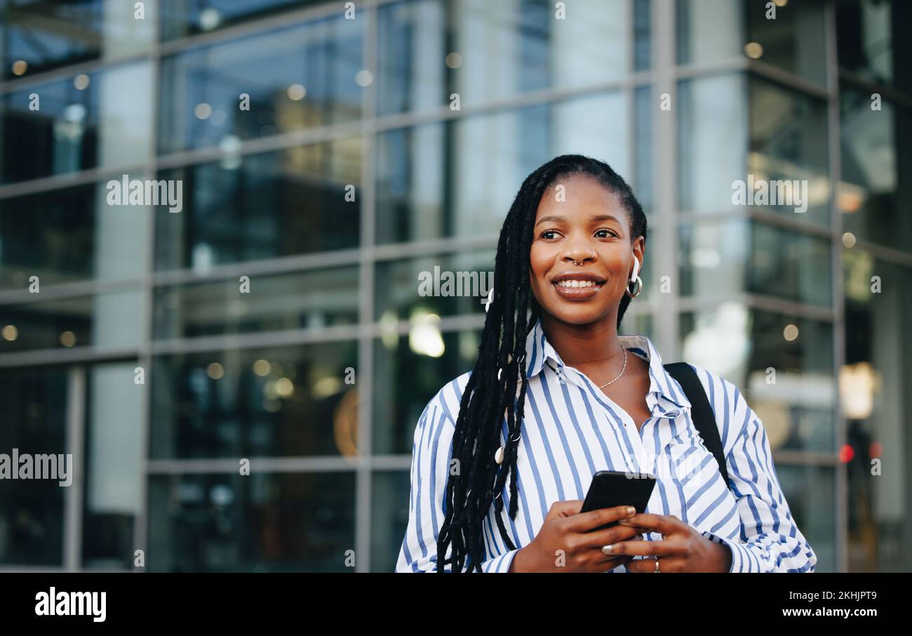 Femme d'affaires pensive tenant un smartphone et portant des écouteurs pendant ses déplacements professionnels en ville. Jeune femme d'affaires noire qui écoute de la musique Banque D'Images