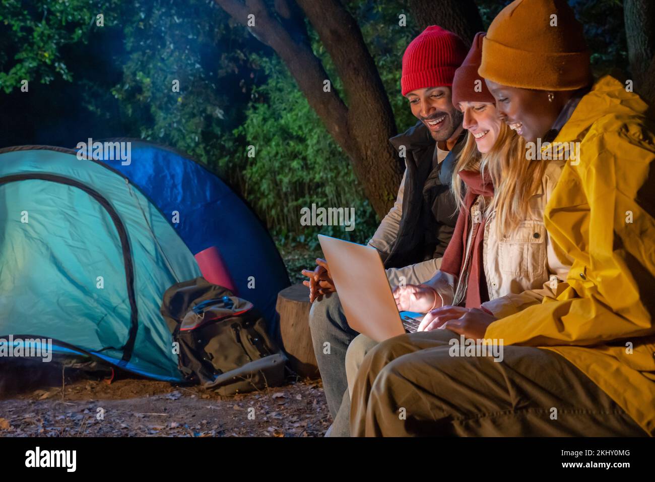 Amis souriants regardant un ordinateur portable tout en campant dans une tente dans la forêt la nuit. Banque D'Images