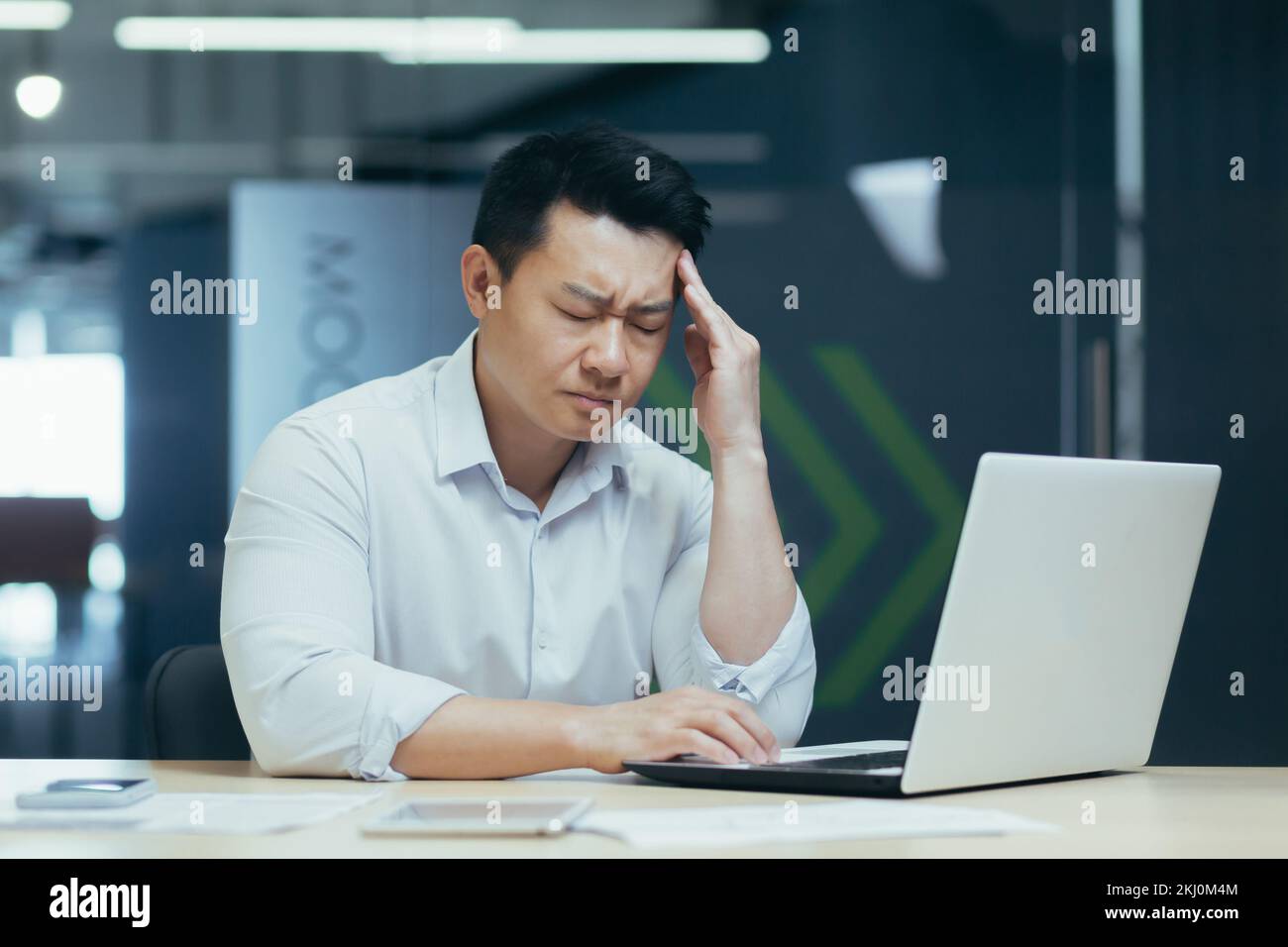 Jeune travailleur de bureau fatigué, indépendant. Assis à un bureau dans le bureau, travaillant sur un ordinateur portable. Il tient la tête, ferme les yeux, ressent un mal de tête sévère, une tension. Banque D'Images