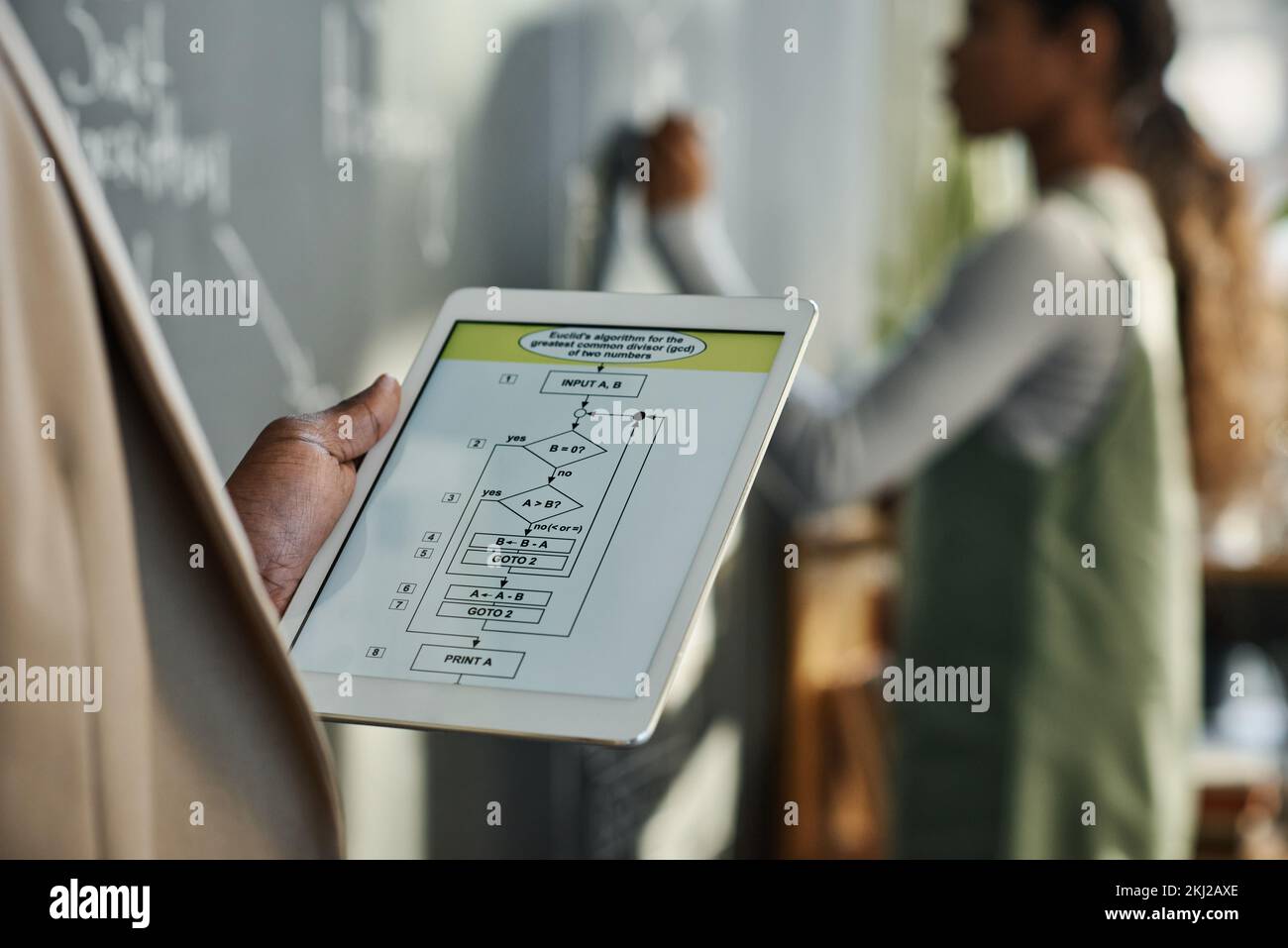 Gros plan de l'enseignant utilisant une tablette numérique avec des graphiques à l'écran avec des enfants écrivant sur le tableau noir en arrière-plan Banque D'Images