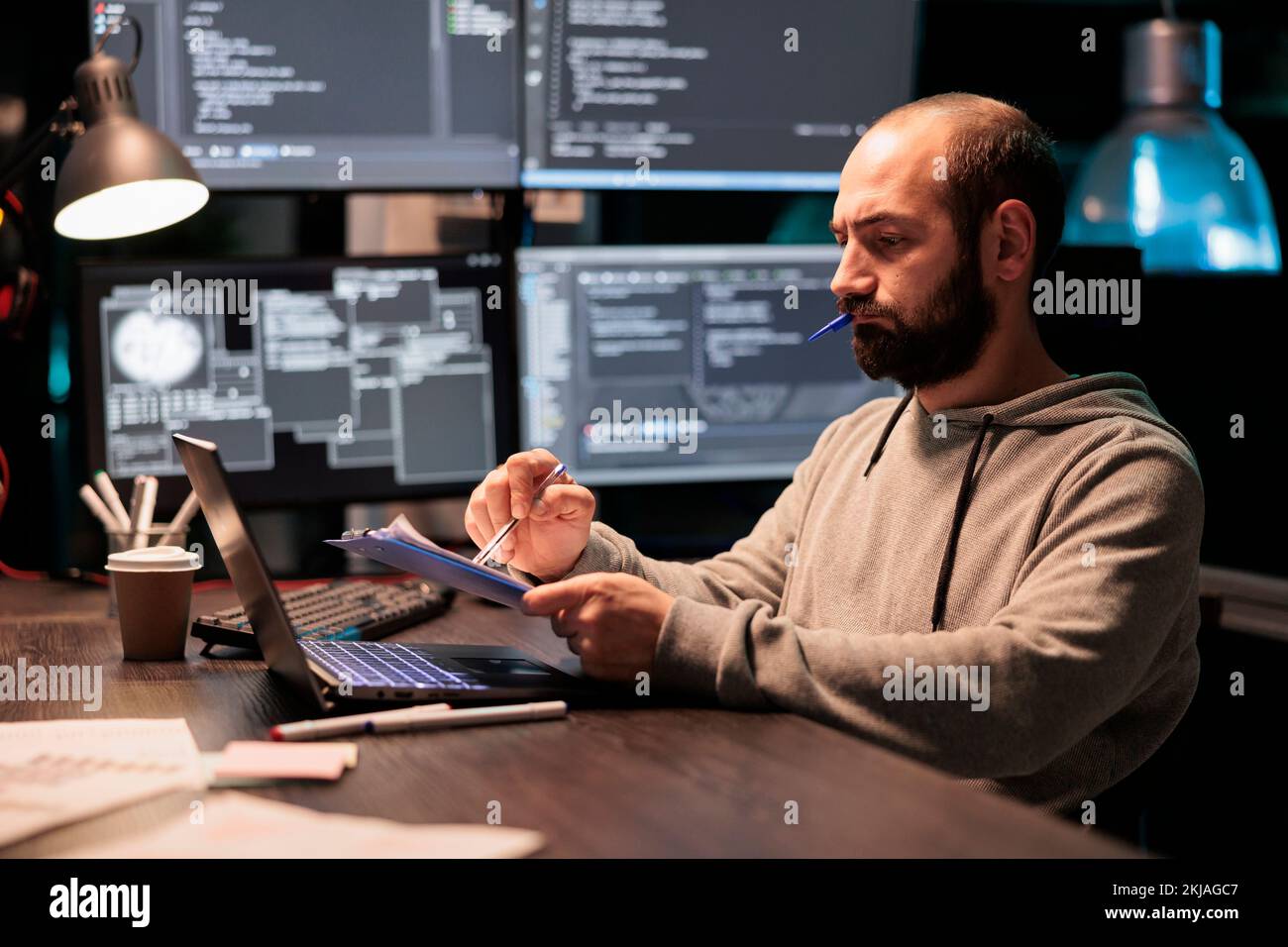 Ingénieur logiciel analysant le code source avec des documents et un ordinateur, prenant des notes et travaillant sur le développement Web. Programmeur masculin comparant les fichiers avec les données du serveur en ligne après les heures de travail. Banque D'Images