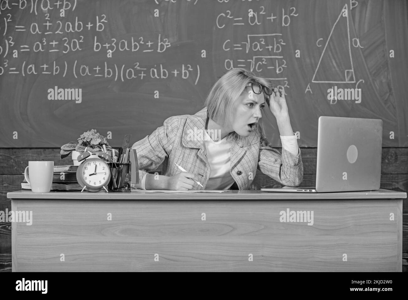 Professeur visage choqué continuer à travailler après les cours. Professeur femme assis table salle de classe tableau noir arrière-plan. Travaillez bien au-delà de la journée scolaire. Toujours Banque D'Images