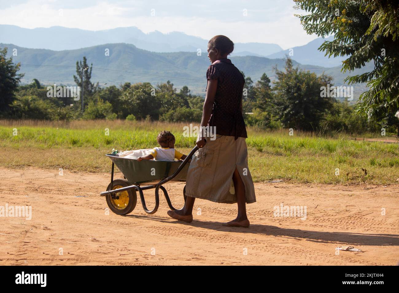 Maman poussant la brouette avec son fils tout-petit à l'intérieur sur une route de terre Banque D'Images
