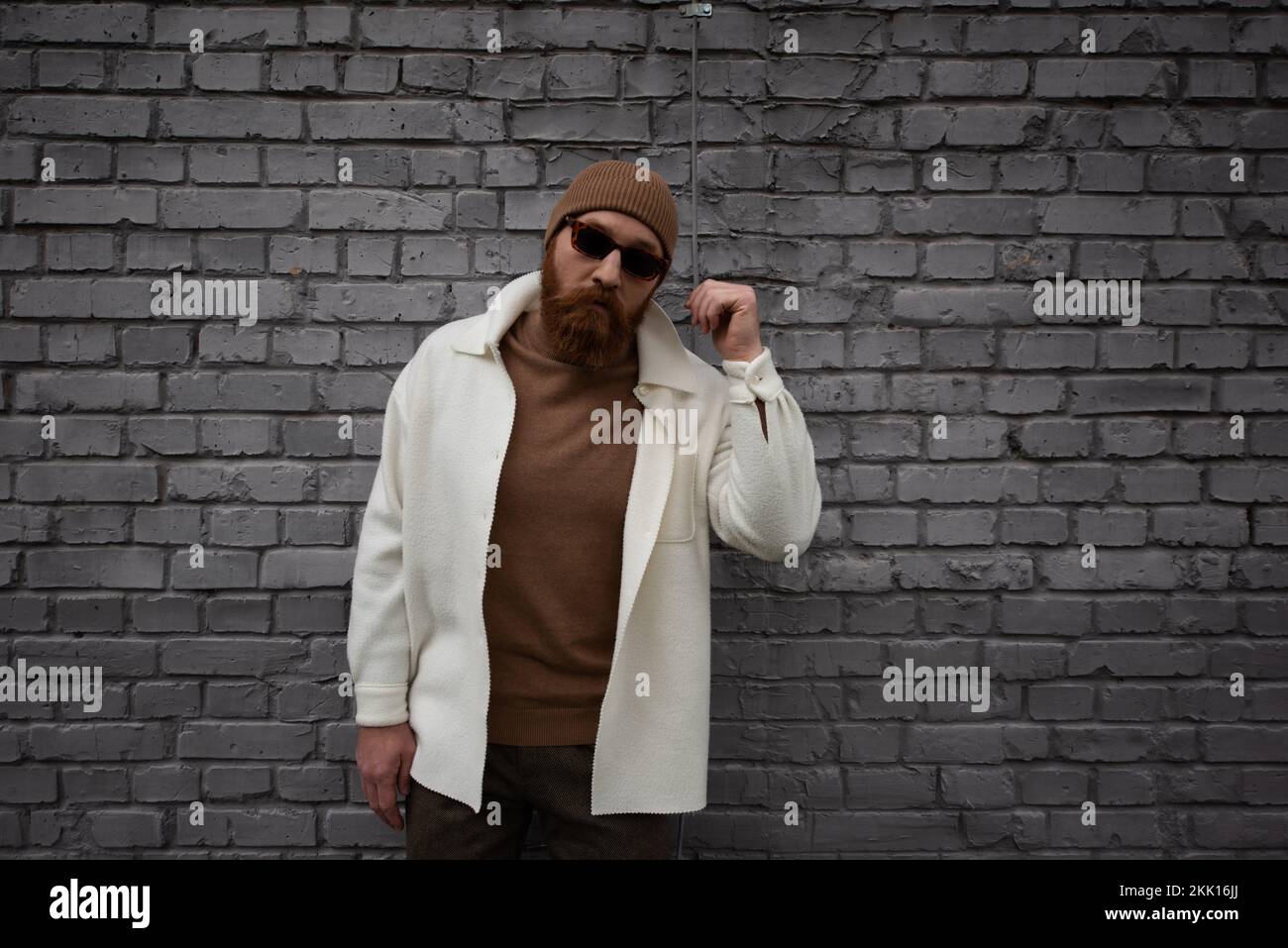 homme élégant et barbu dans bonnet beanie et lunettes de soleil debout près du mur de brique, image de stock Banque D'Images