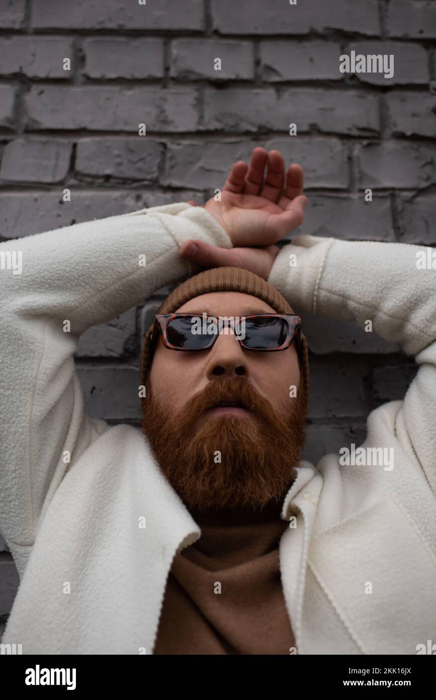 homme élégant et barbu dans bonnet beanie et lunettes de soleil près du mur de brique, image de stock Banque D'Images