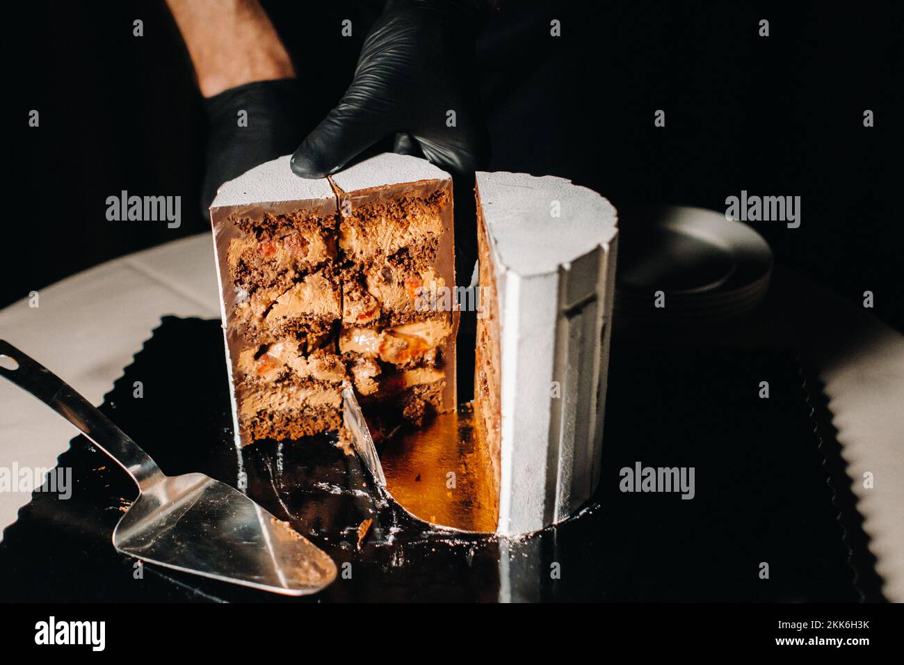 Des gâteaux incroyables. Un chef aux gloved noirs coupe un gâteau de mariage au chocolat. Le gâteau de mariage est délicieux à l'intérieur sur un fond noir.Grand gâteau en blanc Banque D'Images