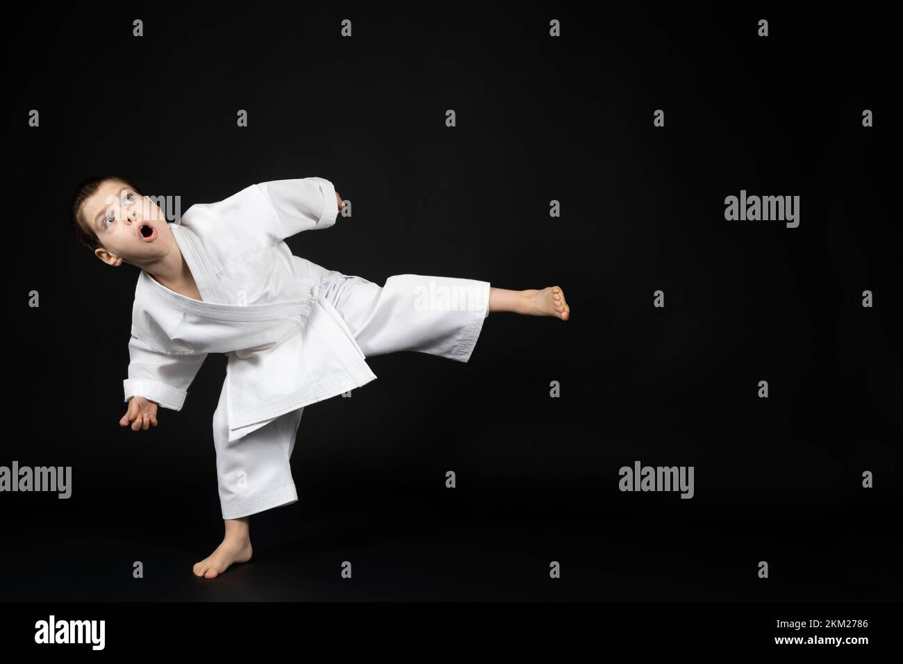 Un petit garçon dans un kimono pratique le karaté sur un fond noir, en avant-pied. Banque D'Images