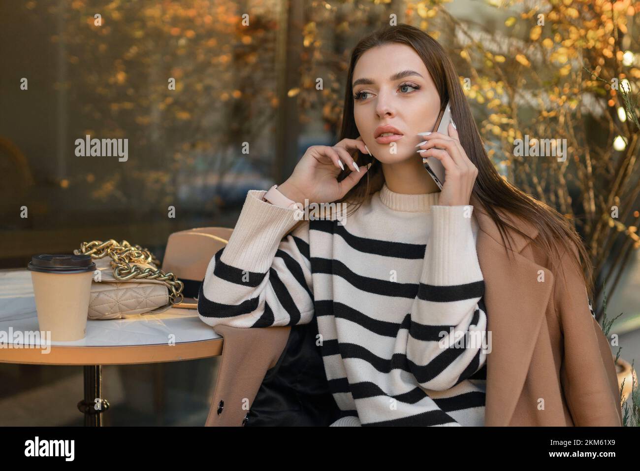 Une brune belle femme caucasienne élégante en vêtements décontractés sur une promenade le long de la rue d'une ville européenne se trouve dans un café et parle au téléphone Banque D'Images