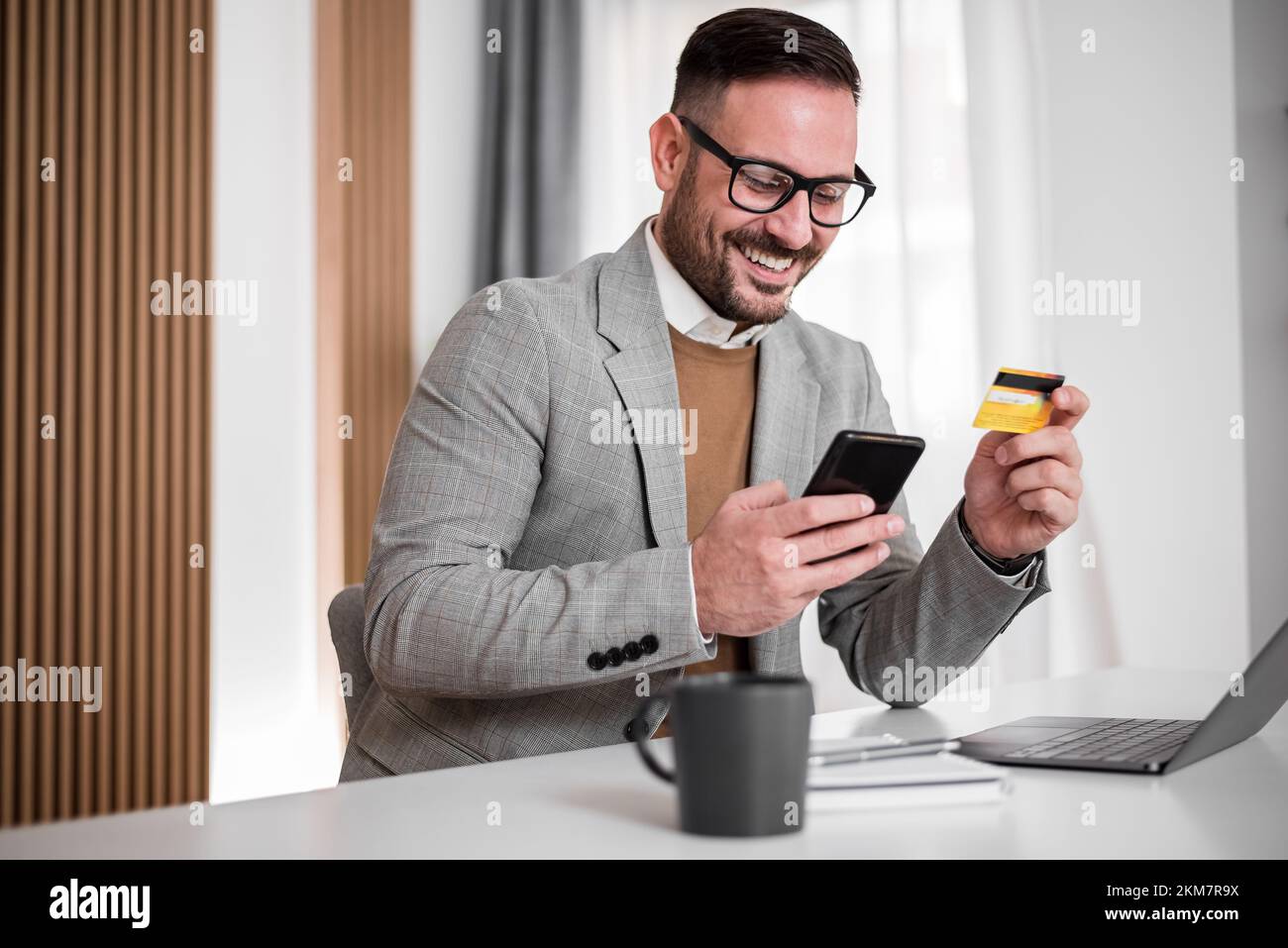 Un cadre pour adultes en costume, en vérifiant son compte bancaire sur son téléphone, souriant tout en tenant sa carte bancaire. Banque D'Images