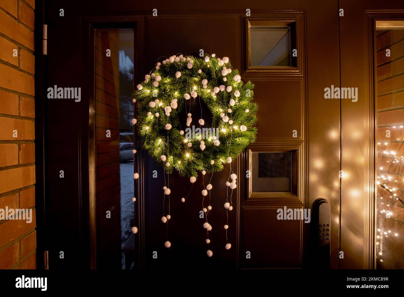Vert épicéa pointes Noël couronne avec lumières chaudes et pompons en feutre blanc sur la porte d'entrée en métal brun avec de minuscules fenêtres à l'extérieur. Banque D'Images