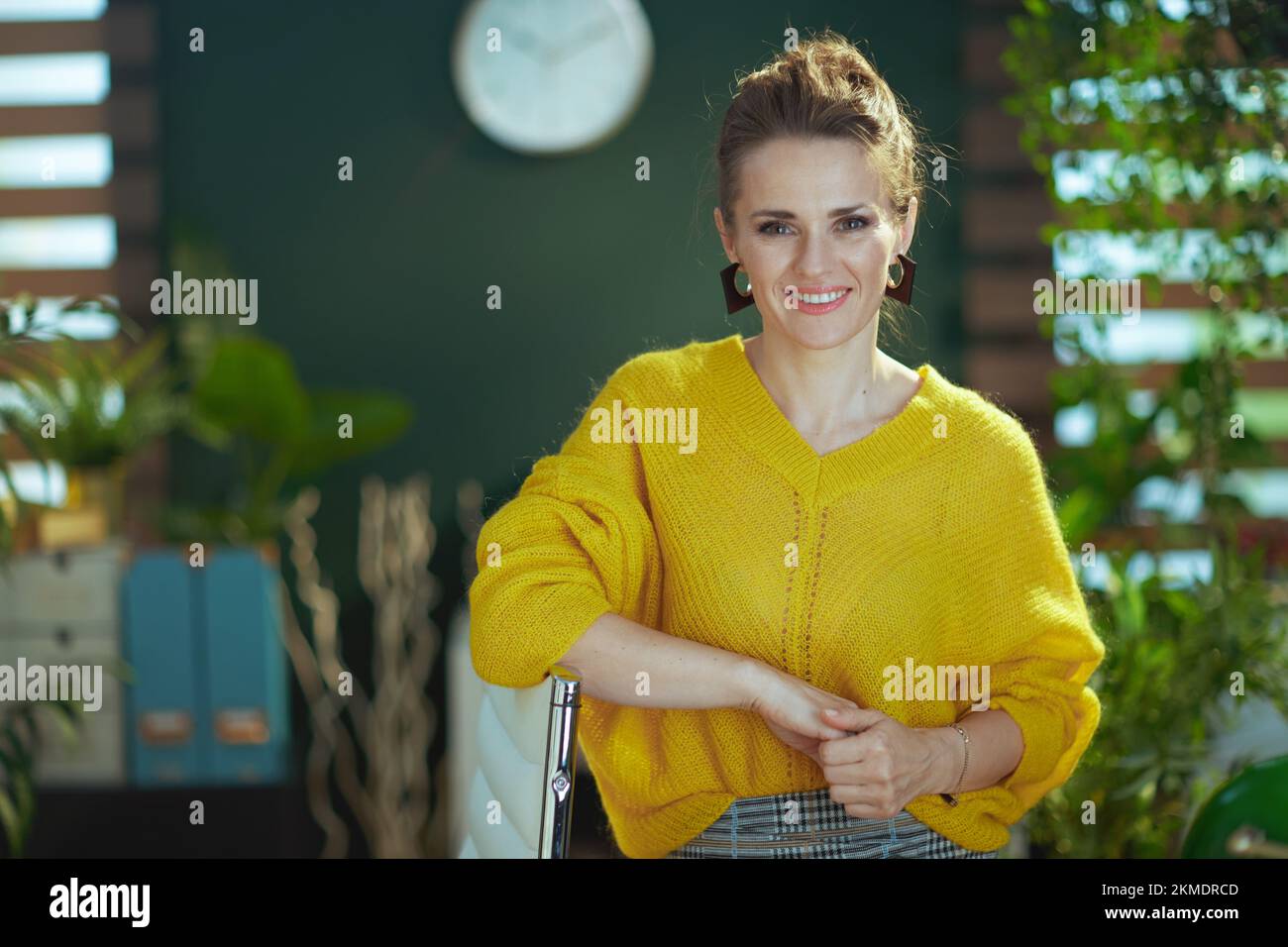 femme souriante et branchée propriétaire d'une petite entreprise dans un chandail jaune debout dans le bureau vert moderne. Banque D'Images